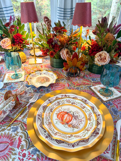 Turkey & Gourds: Thanksgiving Table for 16 set on a patterned tablecloth with gold chargers, teal glasses, and floral centerpieces with candles, featuring paper plates decorated with pumpkin illustrations.