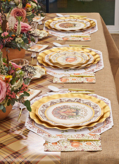 Create a warm autumn tablescape with the Turkey & Gourds Table Setting Bundle from Sophistiplate LLC, featuring designer disposable plates with gold trim, floral-patterned napkins, elegant gold cutlery, and charming autumn floral centerpieces—all beautifully set atop a rustic burlap and plaid tablecloth.