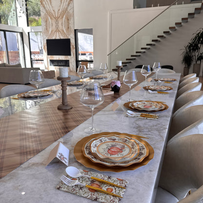 A Turkey & Gourds: Thanksgiving Table for 16 set on a long marble table with pumpkin-illustrated paper plates, gold chargers, patterned napkins, white cutlery, and wine glasses.